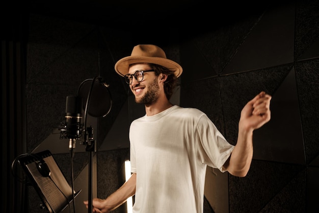 Handsome stylish musician in hat happily dancing recording new song in modern sound studio