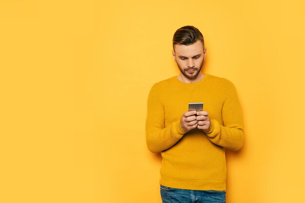 Handsome stylish young beard man in yellow with smart phone in hands is standing over yellow wall