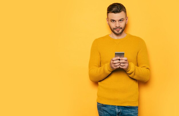 Handsome stylish young beard man in yellow with smart phone in hands is standing over yellow wall