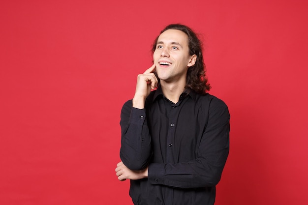 Handsome stylish young curly long haired man in black shirt posing isolated on red background studio portrait. People sincere emotions lifestyle concept. Mock up copy space. Put hand prop up on chin.