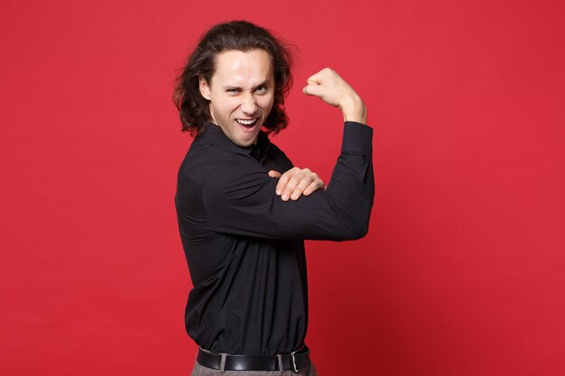 Photo handsome stylish young curly long haired man in black shirt posing isolated on red background studio portrait. people sincere emotions lifestyle concept. mock up copy space. showing biceps, muscles.