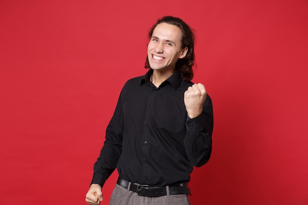 Giovane uomo dai capelli lunghi ricci alla moda bello in camicia nera che posa isolata sul ritratto rosso dello studio del fondo della parete. persone sincere emozioni concetto di stile di vita. mock up spazio di copia. fare il gesto del vincitore.