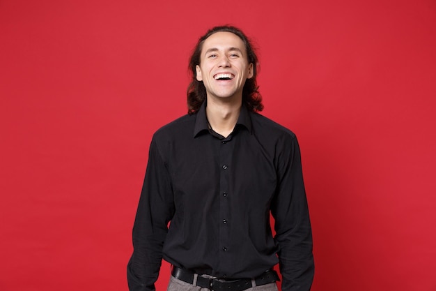 Handsome stylish young curly long haired man in black shirt posing isolated on red wall background studio portrait. people sincere emotions lifestyle concept. mock up copy space. look camera laughing.