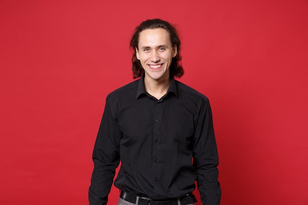 Handsome stylish young curly long haired man in black shirt posing isolated on red wall background studio portrait. people sincere emotions lifestyle concept. mock up copy space. looking camera smile.