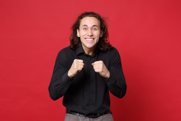 Handsome stylish young curly long haired man in black shirt posing isolated on red wall background studio portrait. People sincere emotions lifestyle concept. Mock up copy space. Looking camera smile.