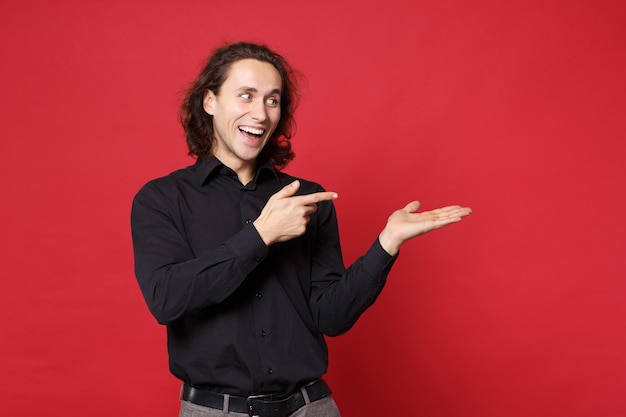 Giovane uomo dai capelli lunghi ricci alla moda bello in camicia nera che posa isolata sul ritratto rosso dello studio del fondo della parete. persone sincere emozioni concetto di stile di vita mock up puntando la mano del dito sullo spazio di copia