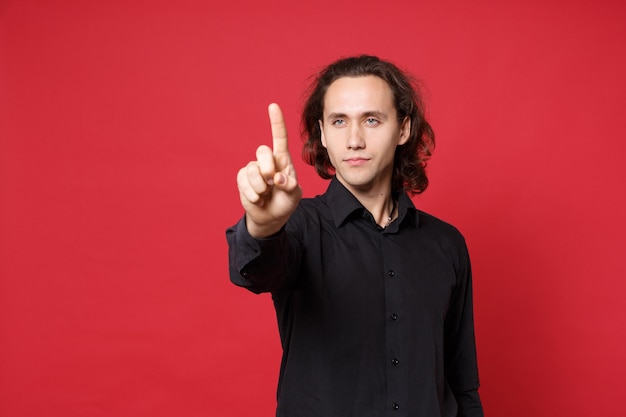 Photo handsome stylish young curly long haired man in black shirt posing isolated on red wall background studio portrait. people sincere emotions lifestyle concept mock up pointing finger hand on copy space