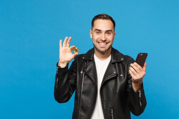 Handsome stylish young unshaven man in black jacket white t-shirt hold in hand cellphone bitcoin currency isolated on blue wall background studio portrait. People lifestyle concept. Mock up copy space