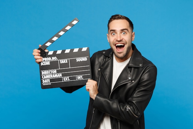 Handsome stylish young unshaven man in black jacket white t-shirt hold in hand film making clapperboard isolated on blue wall background studio portrait. People lifestyle concept. Mock up copy space.