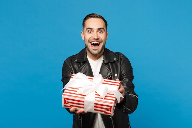 Handsome stylish young unshaven man in black leather jacket white t-shirt hold gift box isolated on blue wall background studio portrait. People sincere emotions lifestyle concept. Mock up copy space.