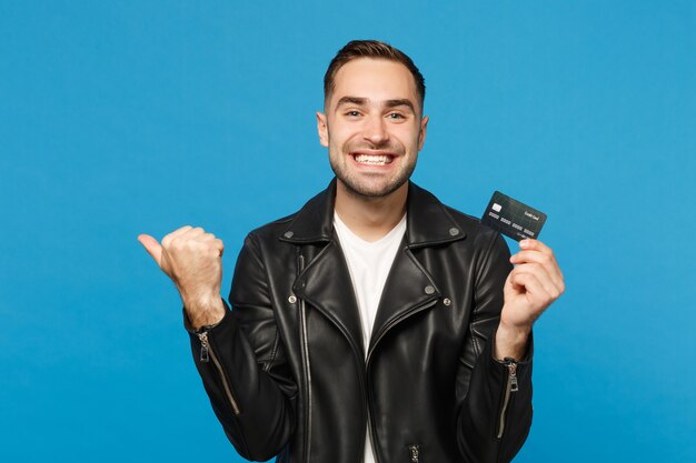 검은 가죽 재킷 흰색 티셔츠를 입은 잘생긴 세련된 젊은 남자는 파란색 벽 배경 스튜디오 초상화에 격리된 신용 은행 카드를 손에 들고 있습니다. 사람들이 라이프 스타일 개념입니다. 복사 공간을 비웃습니다.