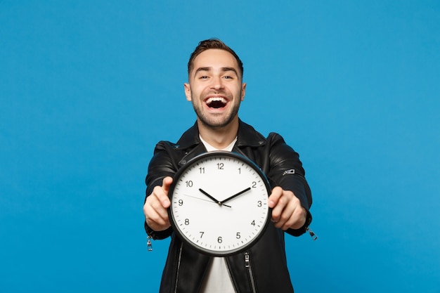 Handsome stylish young unshaven man in black leather jacket white t-shirt holding round clock isolated on blue wall background studio portrait. People lifestyle concept. Hurry up. Mock up copy space.