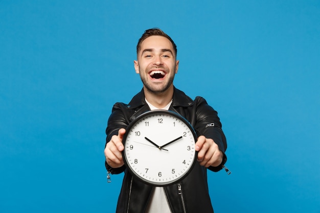 Bello elegante giovane uomo con la barba lunga in giacca di pelle nera t-shirt bianca che tiene orologio rotondo isolato su parete blu sfondo ritratto in studio. concetto di stile di vita della gente. sbrigati. mock up copia spazio.