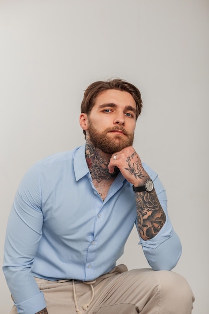 ハンサムな成功したビジネスの流行に敏感な男の入れ墨とファッションの青いシャツにひげを持つ白い背景の上に座っています。