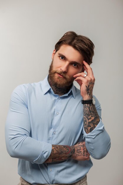 Handsome successful businessman man with a tattoo and a beard in a fashionable blue shirt stands and thinks on a white background in the studio