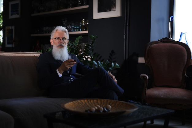 Handsome and successful businessman with stylish bearded in suit holding glass of whiskey while sitting on the sofa in office