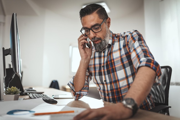 Handsome successful entrepreneur is talking on a cellphone while working in an office and checking documents.