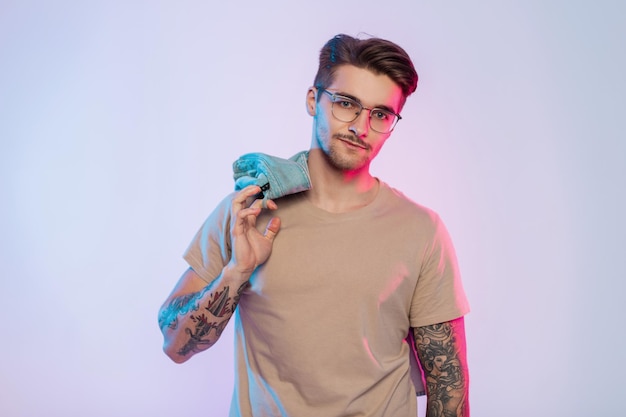 Handsome successful hipster man model with hair and vintage glasses in fashionable casual clothes with a beige Tshirt and jeans shirt with a tattoo on his arms in a studio with pink creative light