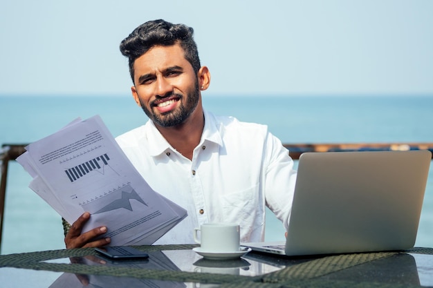 Handsome and successful indian man in a stylish welldressed freelancer working with a laptop on the beachfreelance and remote workbusinessman student in a summer cafe on the shore of india ocean