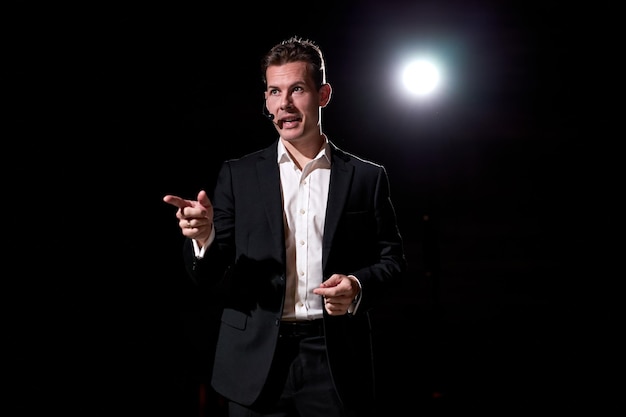 Handsome successful motivational speaker, talking about\
happiness, self, success, empowerment, efficiency, health, and how\
to be more productive. large conference hall with light in\
background