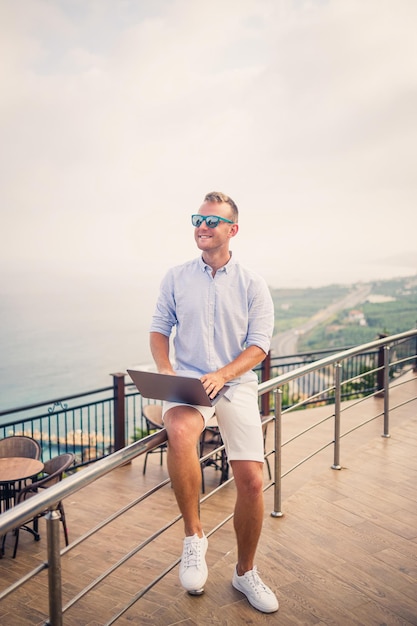 Handsome successful young male businessman working with laptop looks at the Mediterranean Sea He is wearing a shirt and white shorts Remote work on vacation Vacation concept