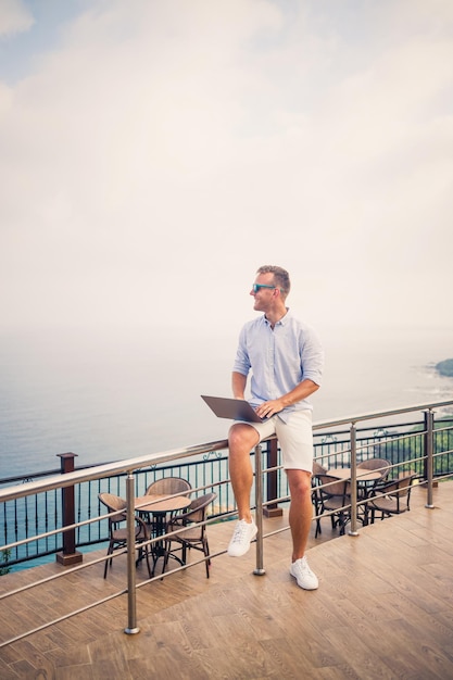 Handsome successful young male businessman working with laptop looks at the Mediterranean Sea He is wearing a shirt and white shorts Remote work on vacation Vacation concept