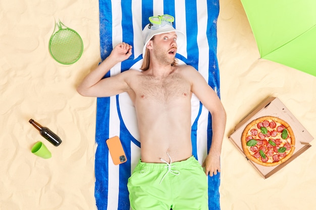 Handsome surprised redheadman wears sunhat and shorts spends leisure time at beach eats delicious pizza drinks beer has lazy day spends summer vacation at seaside. People and recreation concept