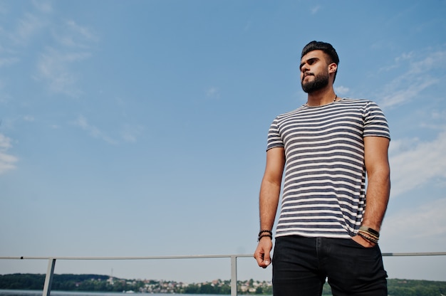 Handsome tall arabian beard man model at stripped shirt posed outdoor against sky. Fashionable arab guy.