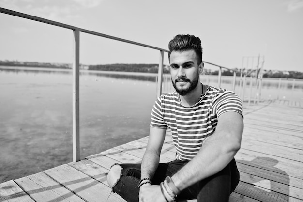 Handsome tall arabian beard man model at stripped shirt posed outdoor on pier of lake Fashionable arab guy