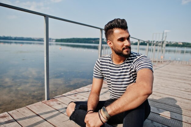 Modello di uomo bello e alto con barba araba con camicia spogliata posata all'aperto sul molo del lago ragazzo arabo alla moda