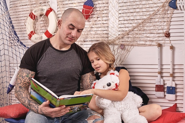 Handsome tattoed man is spending time with his little cute daughter reading fairytales while daughte...