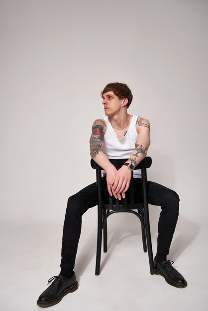 스튜디오의 밝은 배경에 대해 의자에 앉아 있는 흰색 티셔츠를 입은 잘생긴 문신을 한 젊은 남자