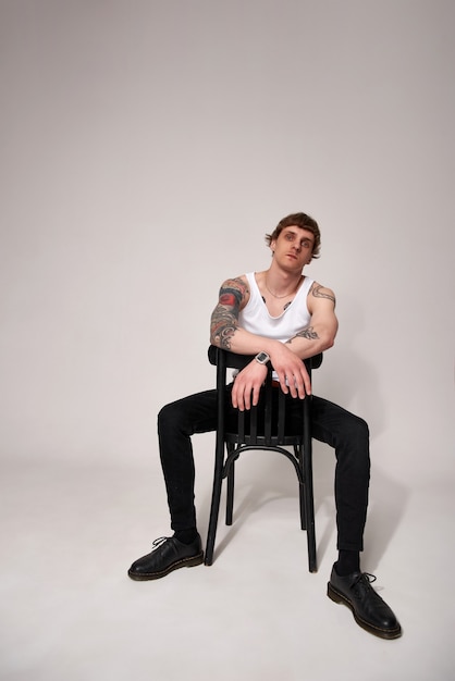 스튜디오의 밝은 배경에 대해 의자에 앉아 있는 흰색 티셔츠를 입은 잘생긴 문신을 한 젊은 남자