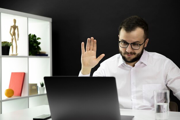 Handsome teacher holds video conference on laptop, raised hand in greeting