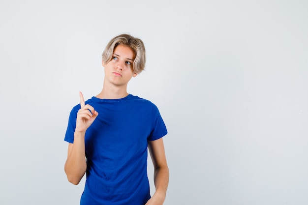 Handsome teen boy pointing up, looking away in blue t-shirt and looking thoughtful , front view.