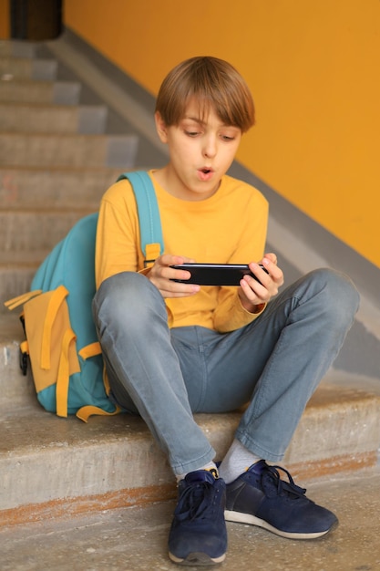 노란색 티셔츠를 입은 잘생긴 10대 소년은 계단에서 전화로 온라인 비디오 게임을 감정적으로 한다