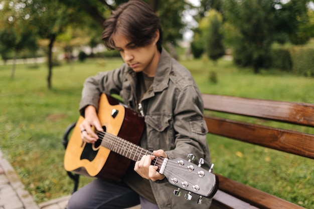 Handsome teenage playing acoustic guitar outdoor in autumn time boy sitting on bench and playing