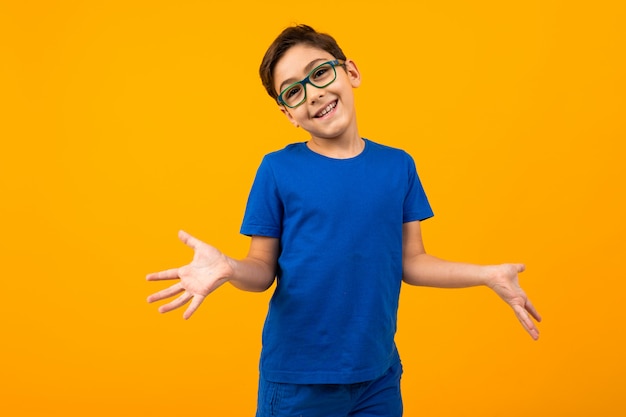 Handsome teenager boy in a blue t-shirt with glasses holds out his hands in different directions on yellow with copy space