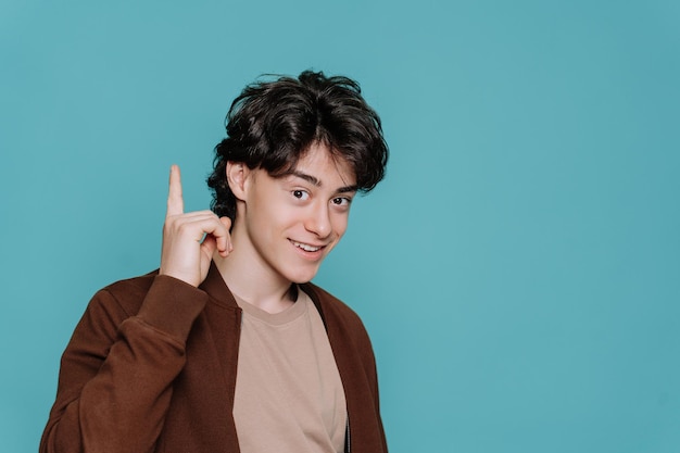 Handsome teenager boy in green brown jacket points by index\
finger up at workspace area isolated on plain turquoise background\
studio portrait mockup sale discount idea happy guy looks at\
camera