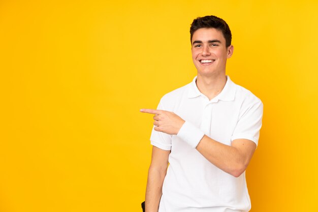 Handsome teenager tennis player man isolated on yellow playing tennis and pointing to the lateral