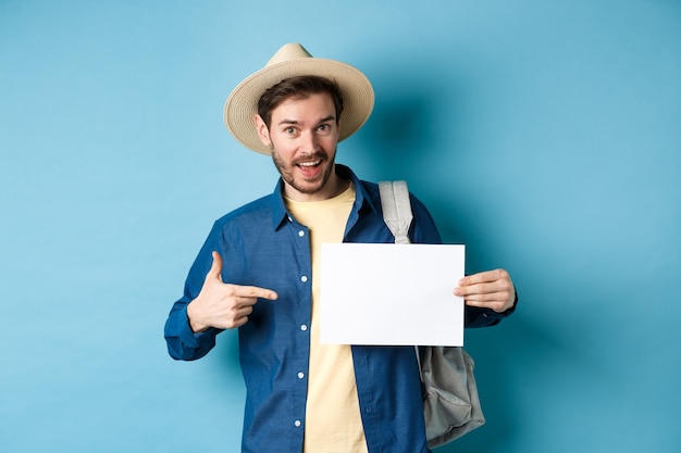 Handsome tourist smiling and pointing at empty piece of paper, wearing straw hat and backpack. Concept of summer vacation.