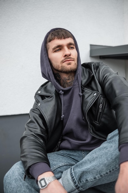 Handsome trendy hipster model guy with hair and a beard in a fashionable leather rock jacket with hoodies and jeans sits and rest near a white building on the street