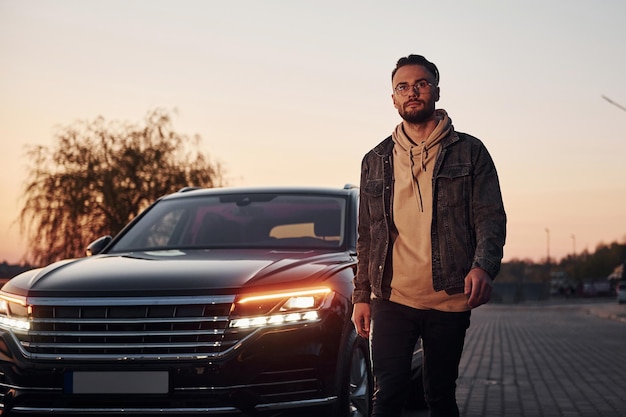 Handsome unshaved man in fashionable clothes walks near his black car at evening time