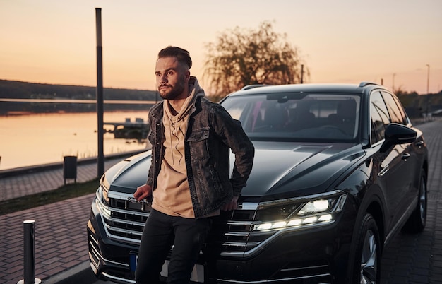 Handsome unshaved man in fashionable clothesleaning on his his black car