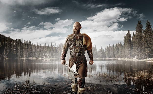 Handsome viking with axes in hands dressed in traditional nordic clothes standing at the lake. Ancient warrior at the river, northern forest