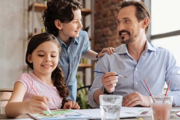 Handsome wavy-haired boy resting his hand on the shoulder of father and talking to him while his little sister painting a watercolor picture