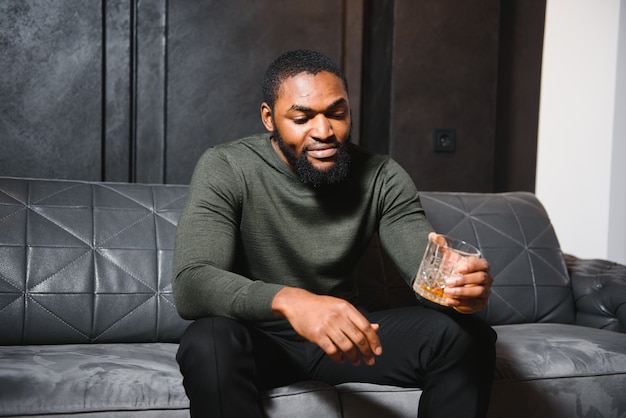 Handsome young african american man in elegant suit with glass of whiskey relaxing on sofa