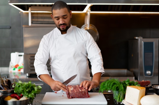 Handsome young African chef standing in professional kitchen in restaurant preparing a meal of meat and cheese vegetables. Portrait of man in cook uniform Cuts meat with a metal knife.