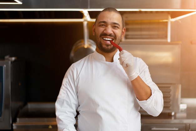 Handsome young African chef standing in professional kitchen in restaurant preparing a meal of meat and cheese vegetables. Portrait of man in cook uniform. Taste red pepper