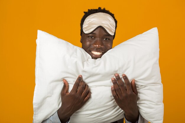 Handsome young african male in a jacket wearing a sleep mask with a pillow in his hands